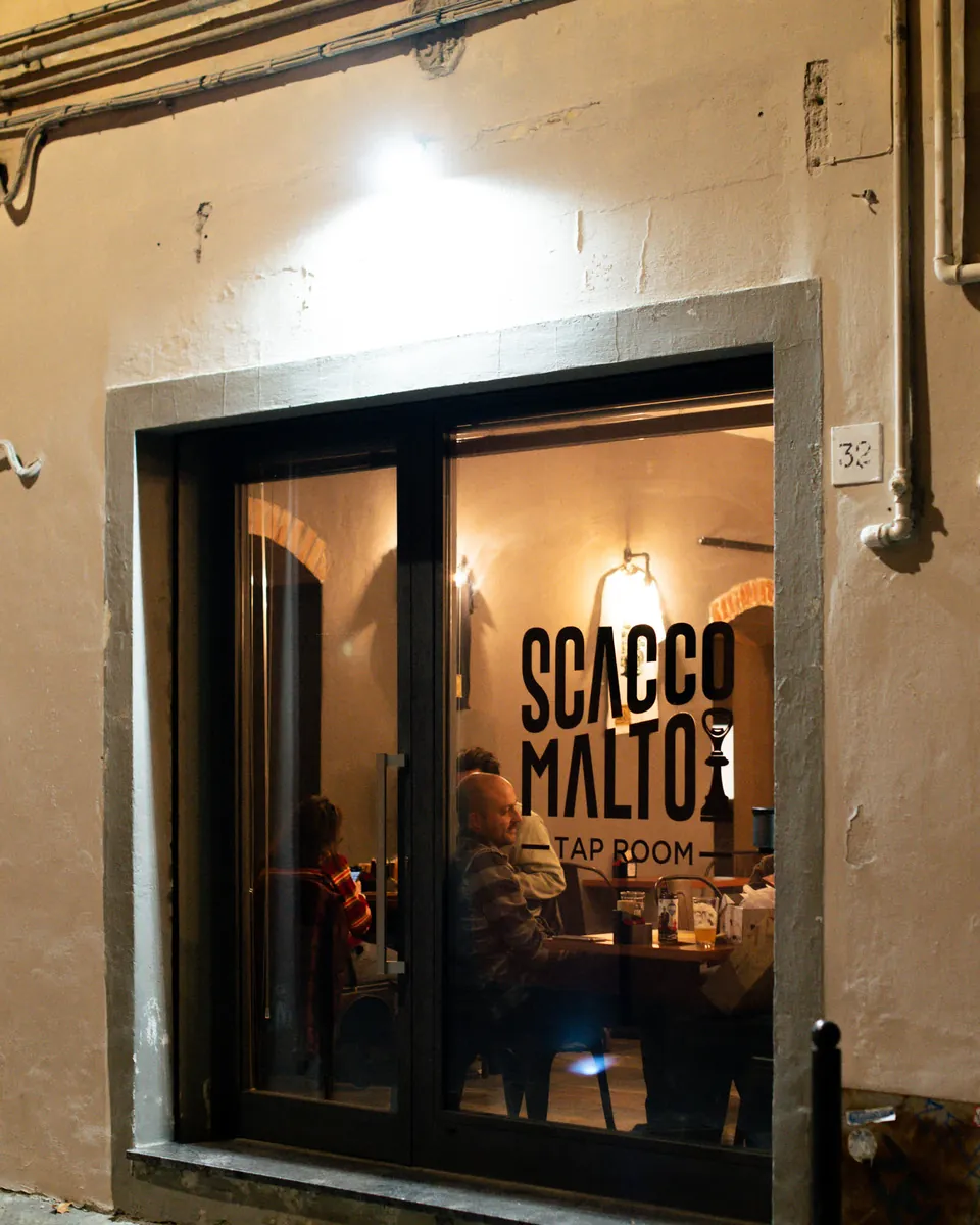 La vetrina di Scaccomalto illuminata di sera, con la gente dentro